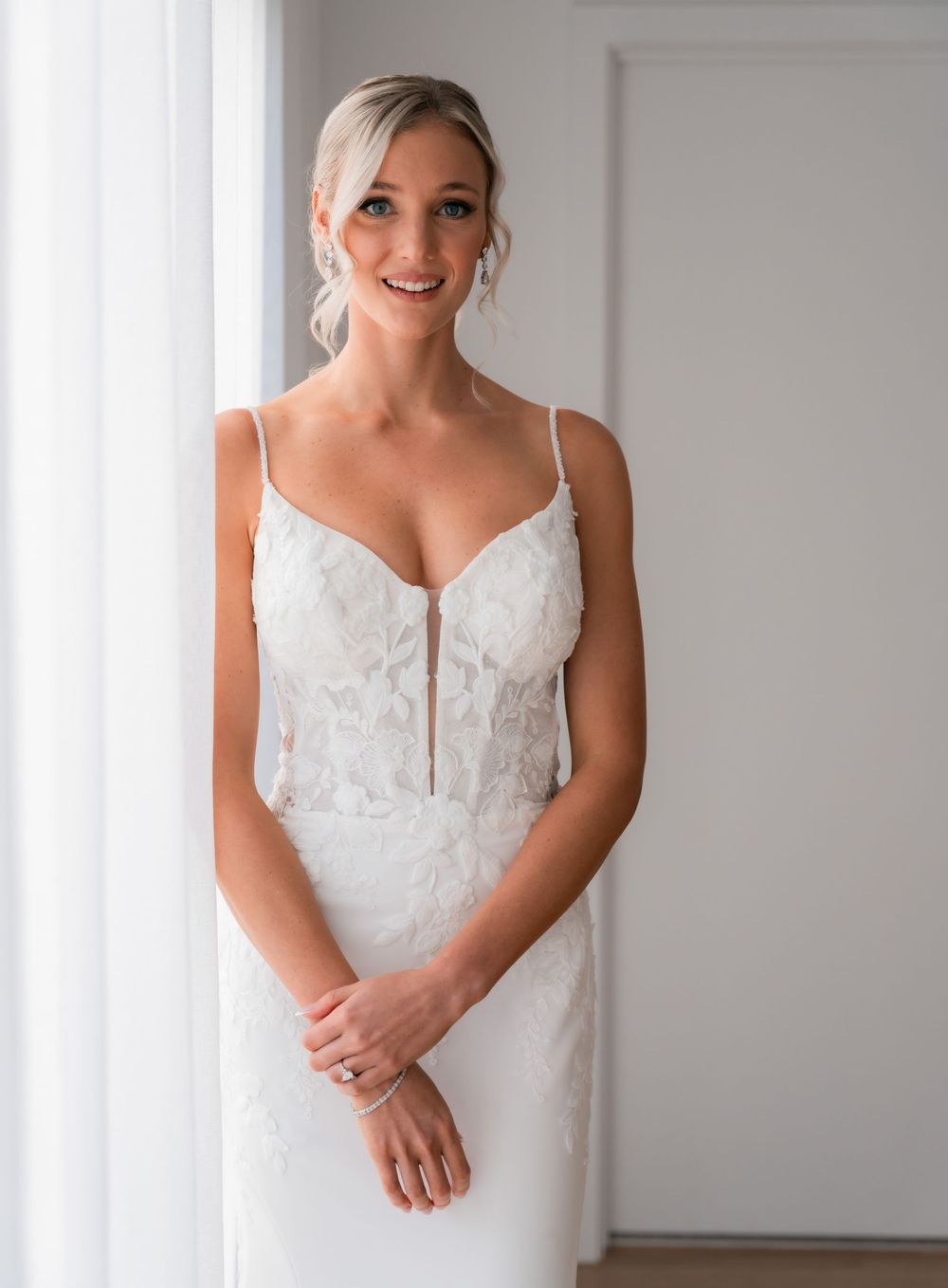 Smiling bride in a white lace spaghetti strap wedding gown posing elegantly near a bright window.