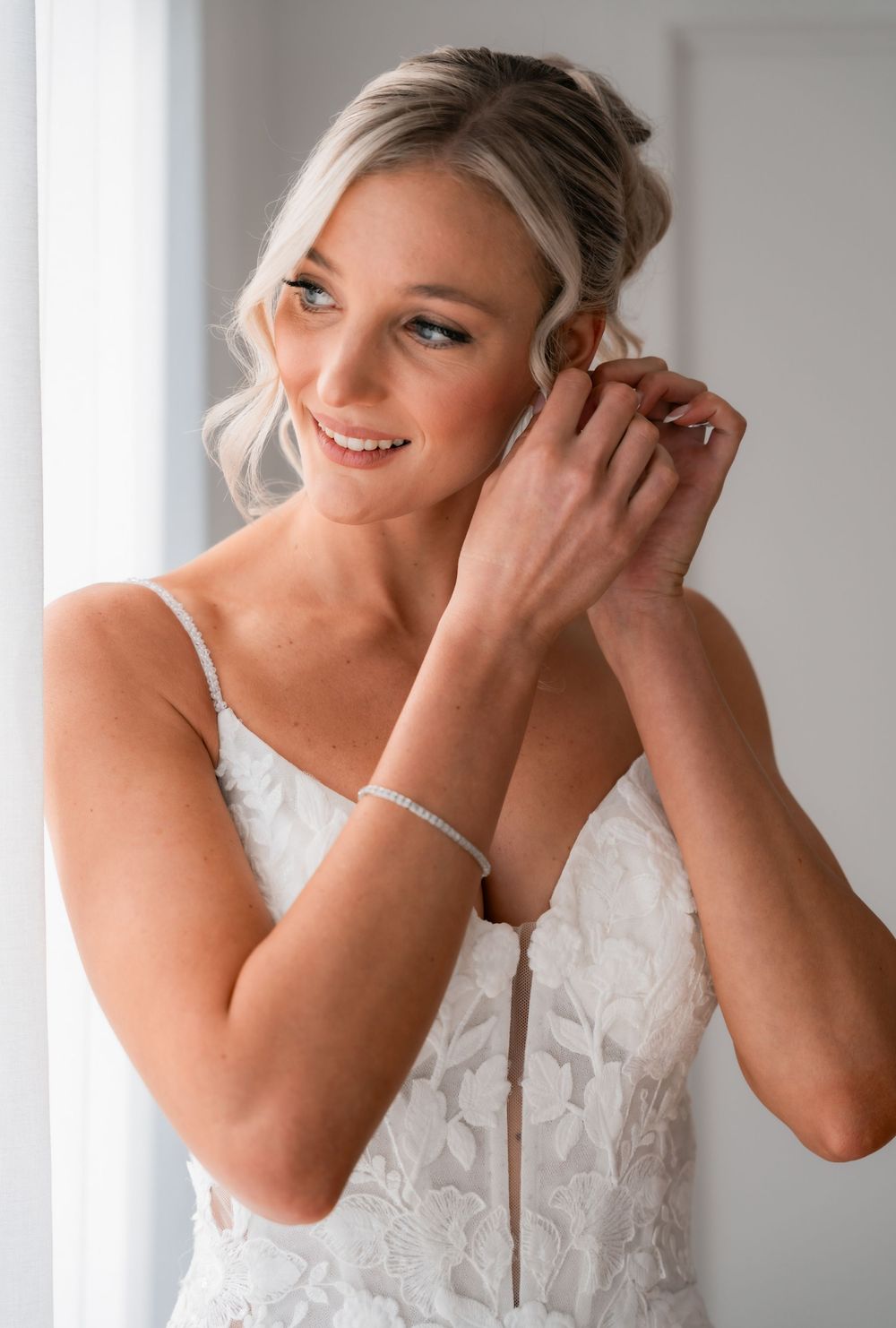 Smiling bride in lace gown adjusting earring, wearing diamond bracelet, soft updo hairstyle, near bright window.