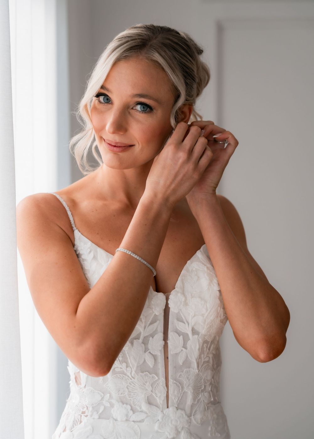 Bride in lace wedding gown adjusting earring near window, wearing delicate bracelet with elegant updo hairstyle.