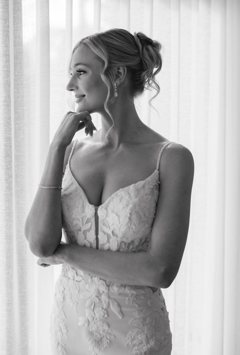 Black and white photo of a smiling bride in a lace wedding gown with spaghetti straps, elegant updo, gazing out a window.
