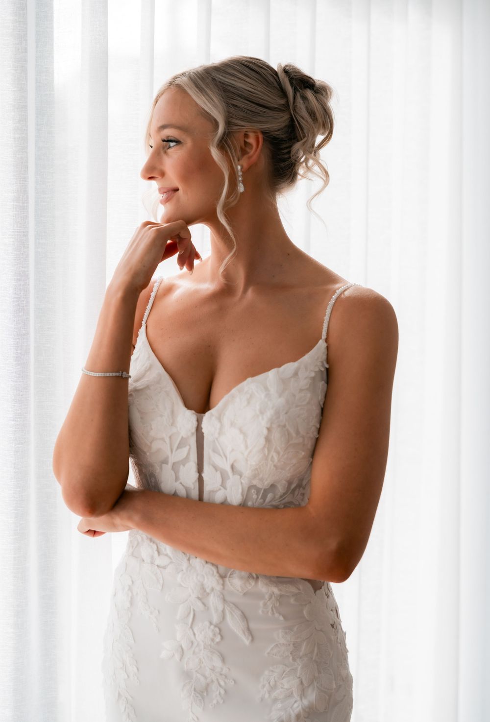 Bride in a white lace spaghetti strap wedding gown with updo hairstyle and drop earrings gazing softly by a window.