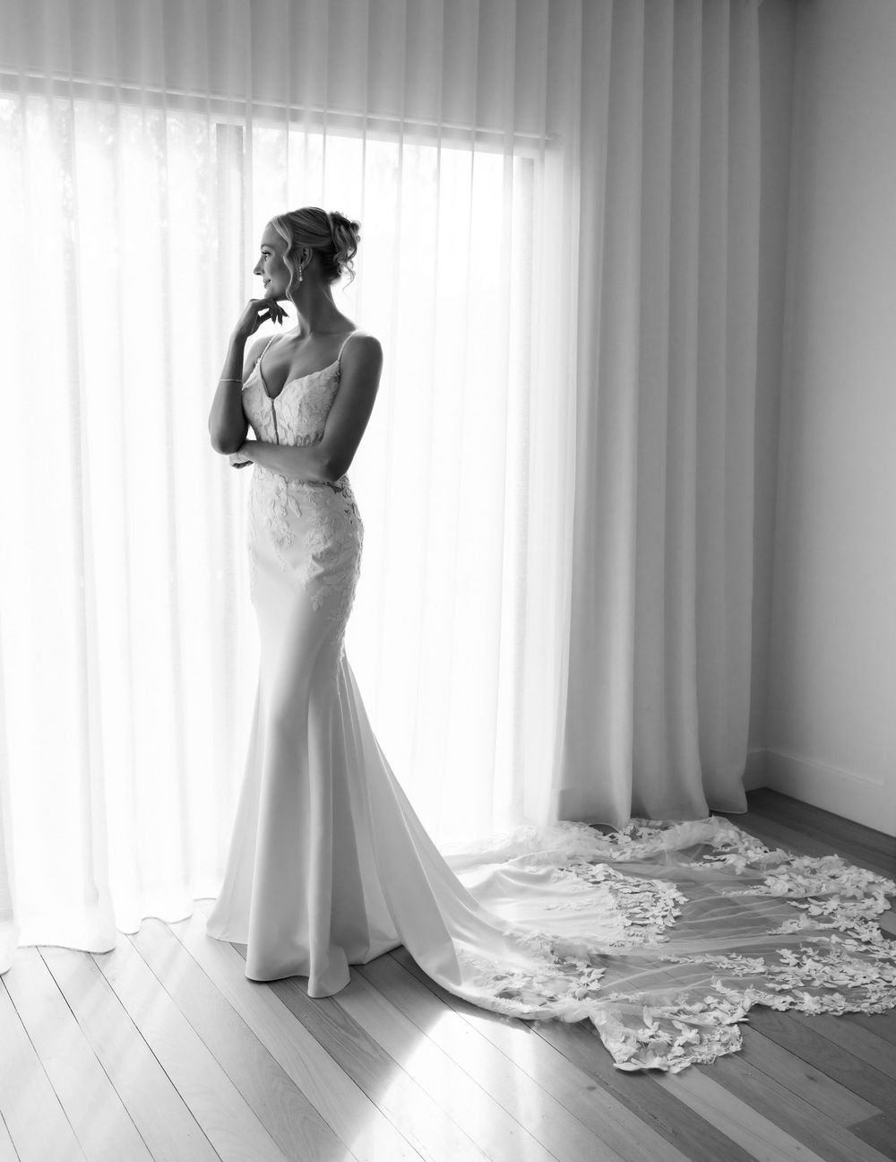 Elegant bride in a fitted mermaid wedding gown with lace train stands by sheer curtains in black and white photo.