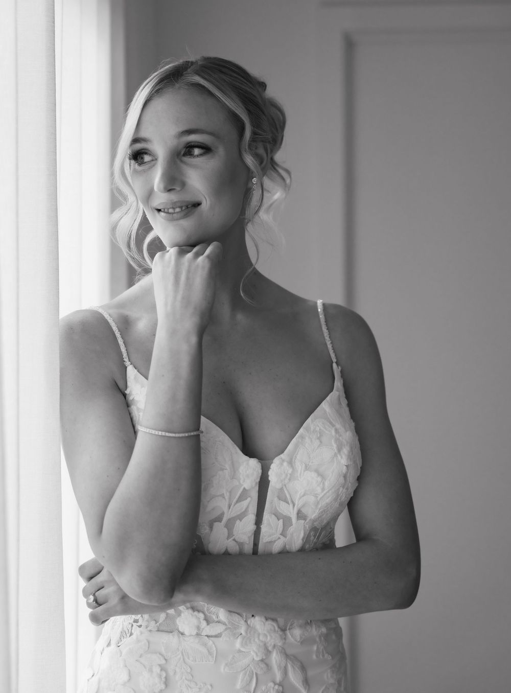 Black and white bridal portrait of a blonde bride in lace gown gazing thoughtfully by a window.