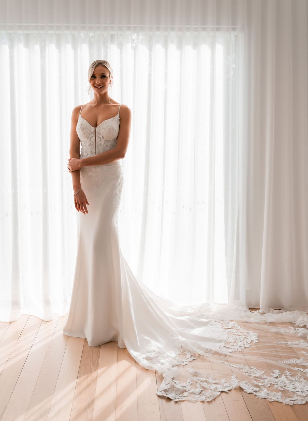 Elegant bride in a fitted white lace bodice wedding gown with a flowing train, standing before sheer curtains.