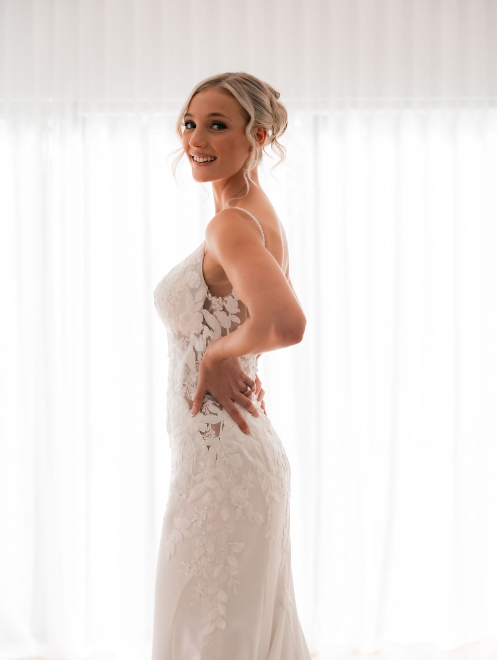 Bride in a fitted white lace wedding gown smiling over her shoulder, posing elegantly in soft natural light.