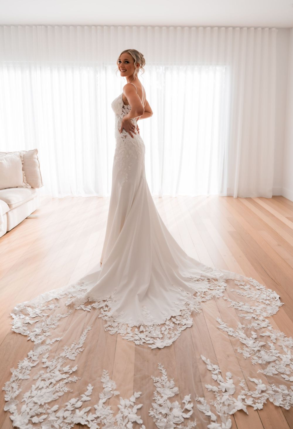 Bride in strapless white wedding gown with floral lace train standing by bright window on hardwood floor.