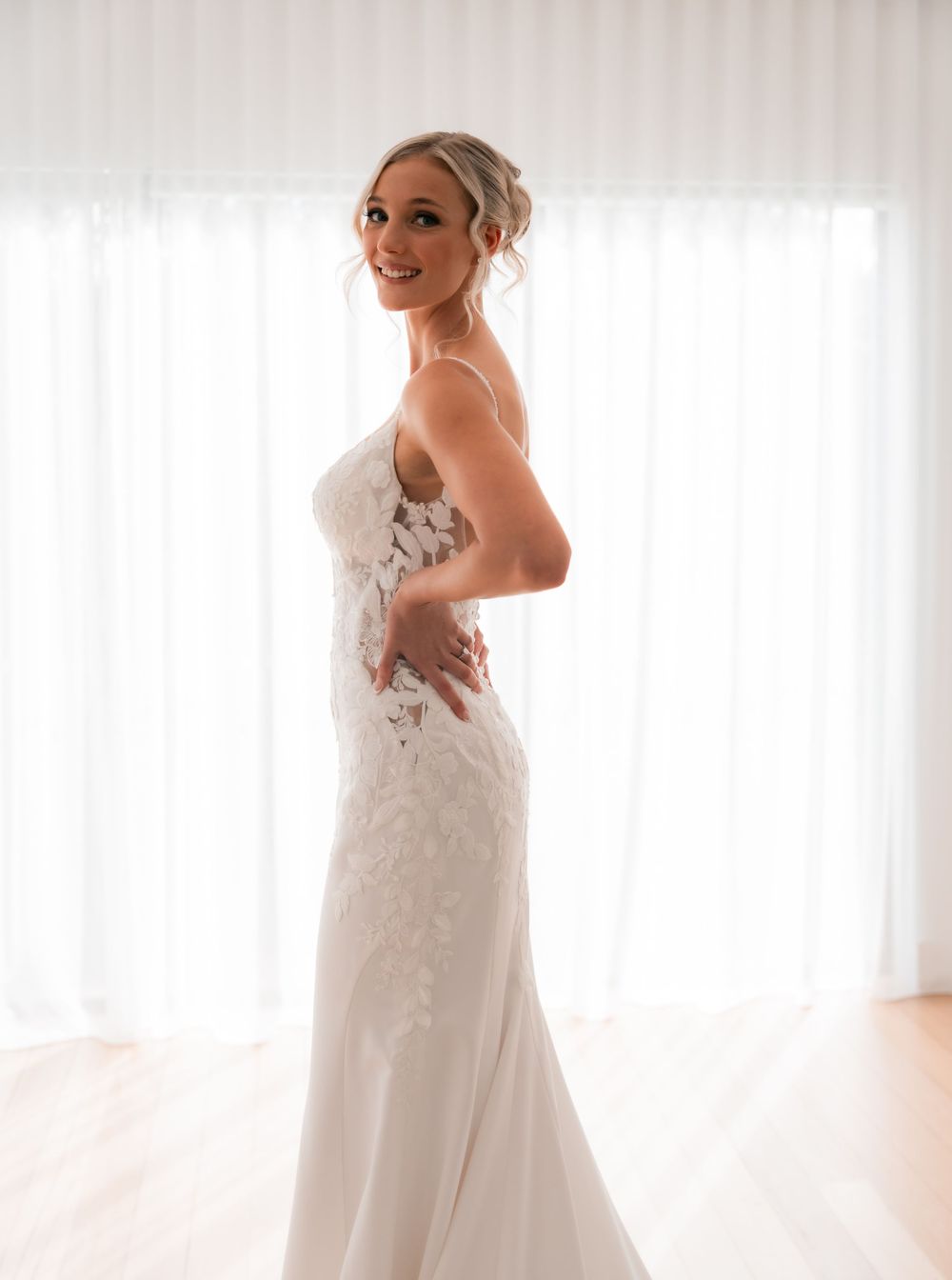 Bride in elegant white lace wedding gown posing sideways, smiling over shoulder in bright room with sheer curtains.