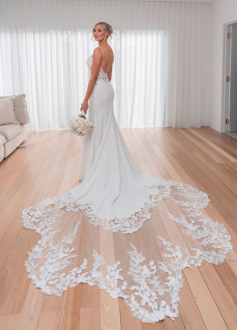 Bride in elegant backless white wedding gown with long lace cathedral train, holding bouquet on hardwood floor.