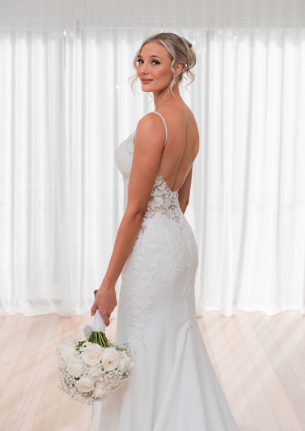 Bride in backless lace wedding gown with spaghetti straps holds white rose bouquet, posing elegantly in bright studio.