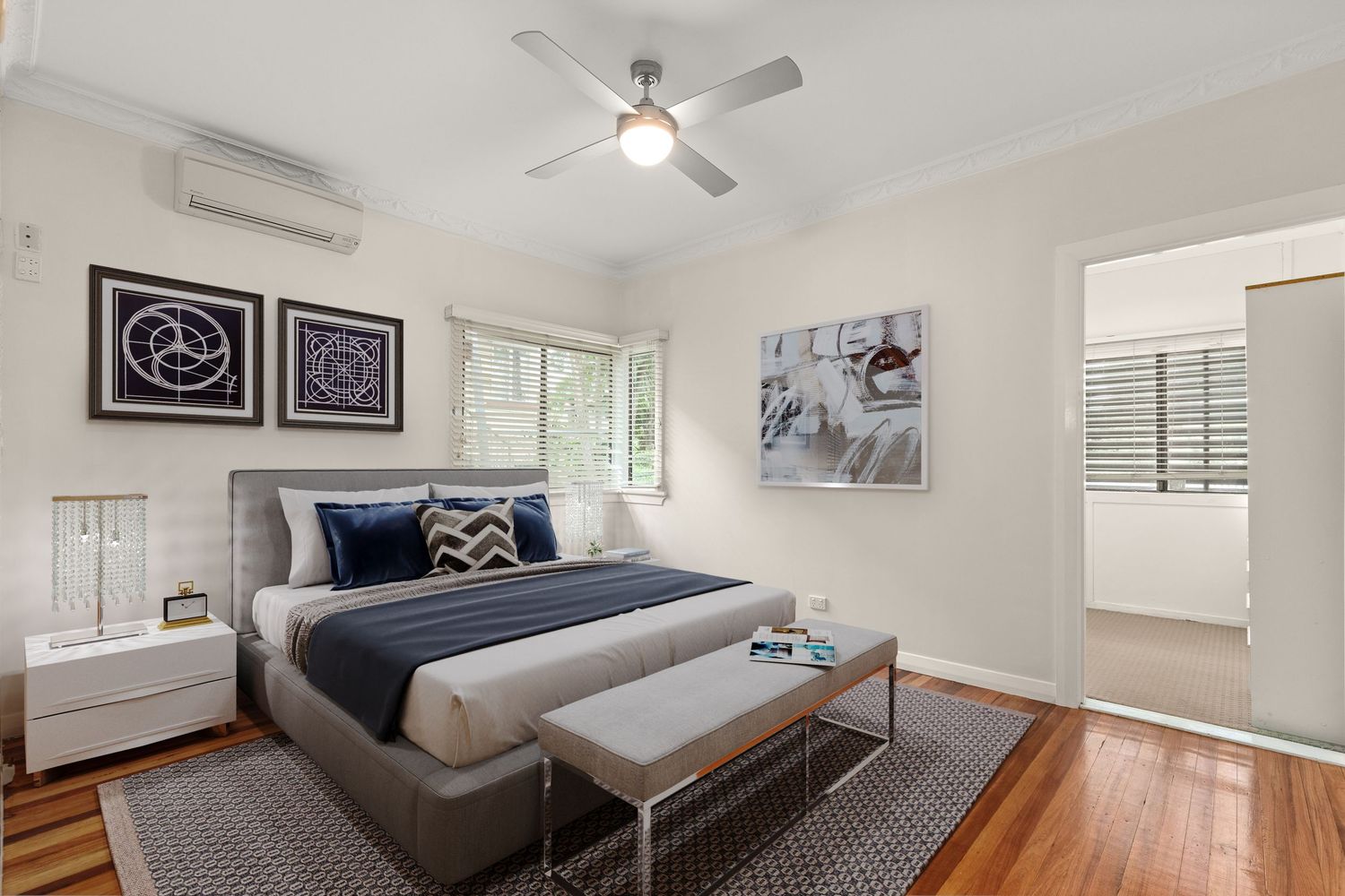 Stylish bedroom with hardwood floors, ceiling fan, split-system AC, blue bedding, and plantation shutters on window.