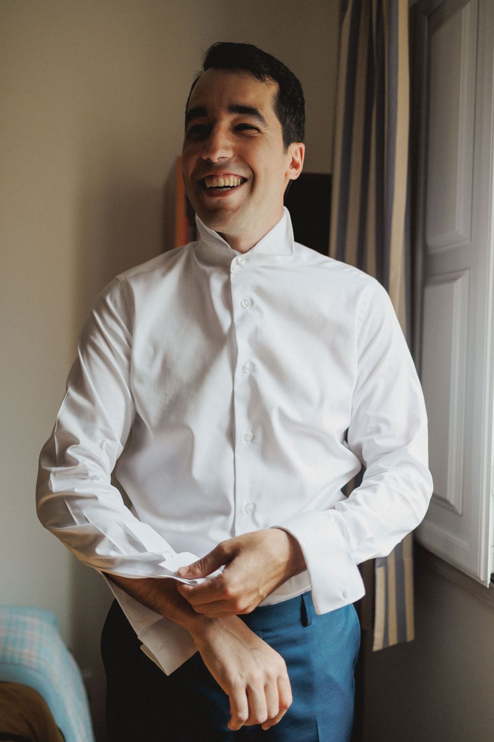 A groom in a white button-up shirt and blue pants laughs joyfully while he's getting ready for a wedding in azeitão