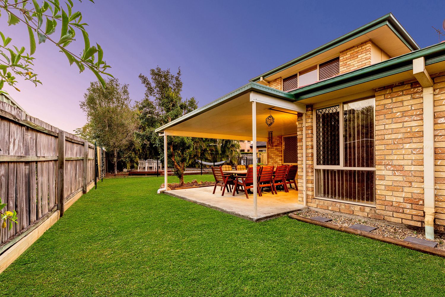 Covered outdoor entertaining area with dining set, green lawn, and timber fence at dusk.