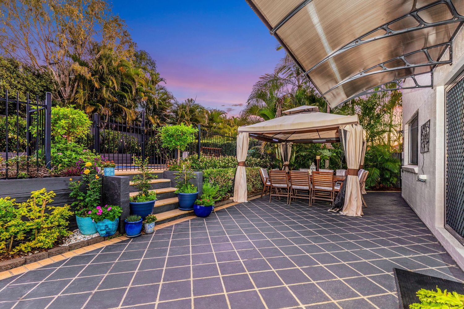 Outdoor patio with tiled flooring, gazebo dining area, garden beds, and pergola at dusk.