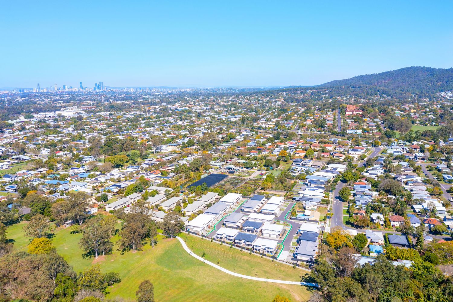 Aerial view of a Brisbane suburb featuring a residential complex, lush parklands, and city skyline in the background.
