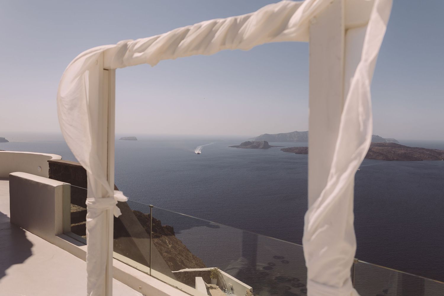 White curtains wrapped round a wooden frame blow in the breeze against a backdrop of Santorini caldera.