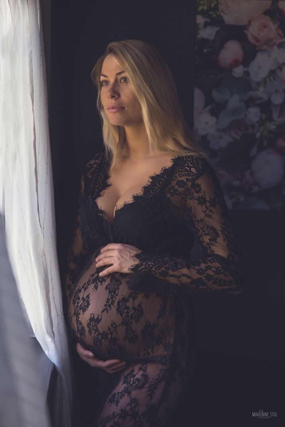 Artistic maternity photo featuring an expectant mother in a black lace gown standing by a window in soft lighting.