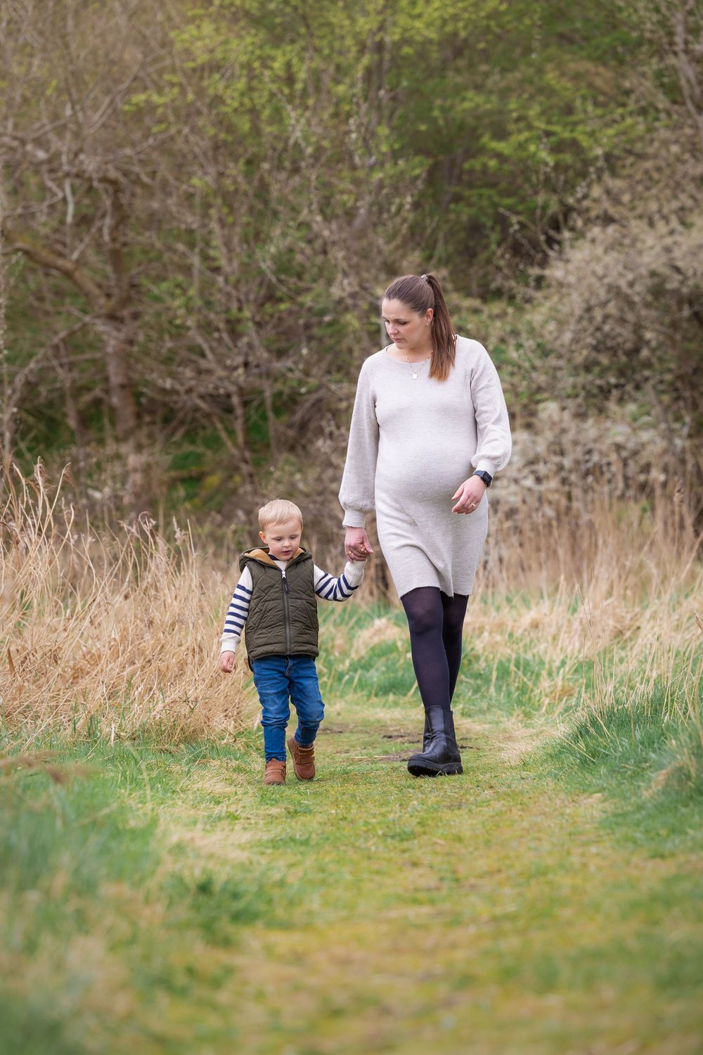 A pregnant woman and small child walking together on a grassy nature path during autumn.