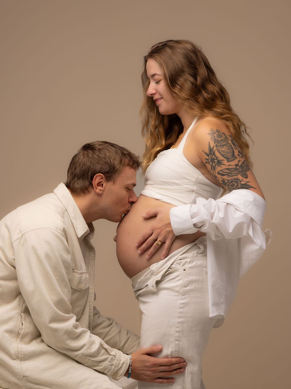 A man kisses a pregnant woman's belly during a maternity photoshoot, both wearing white outfits against a beige background.