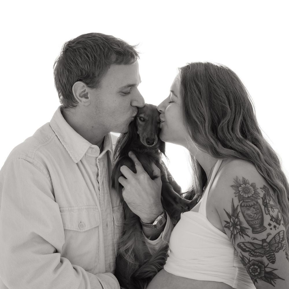 Black and white photo of a couple kissing their small dachshund dog, woman with tattoos, white background.