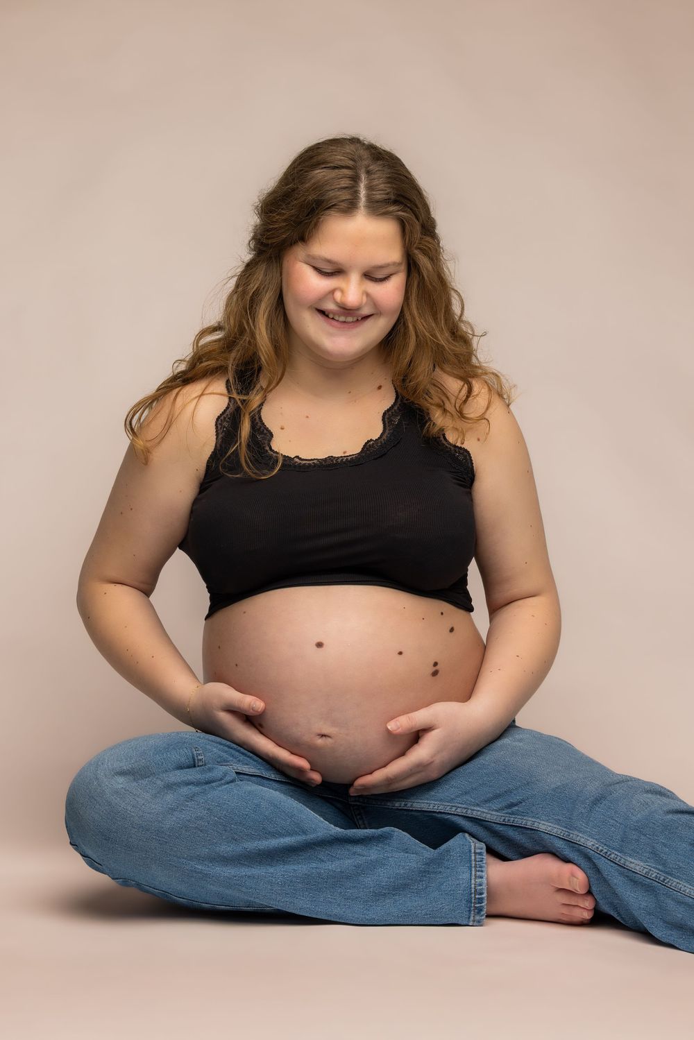 Smiling pregnant woman in black crop top and jeans sitting cross-legged, cradling her bare baby bump.