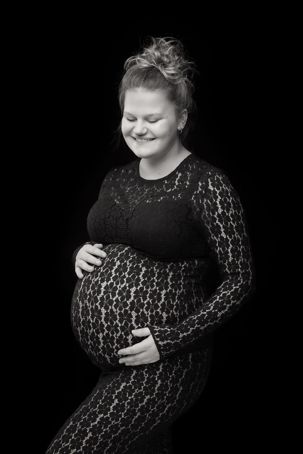 Black and white portrait of a smiling pregnant woman in lace dress cradling her baby bump against dark background.