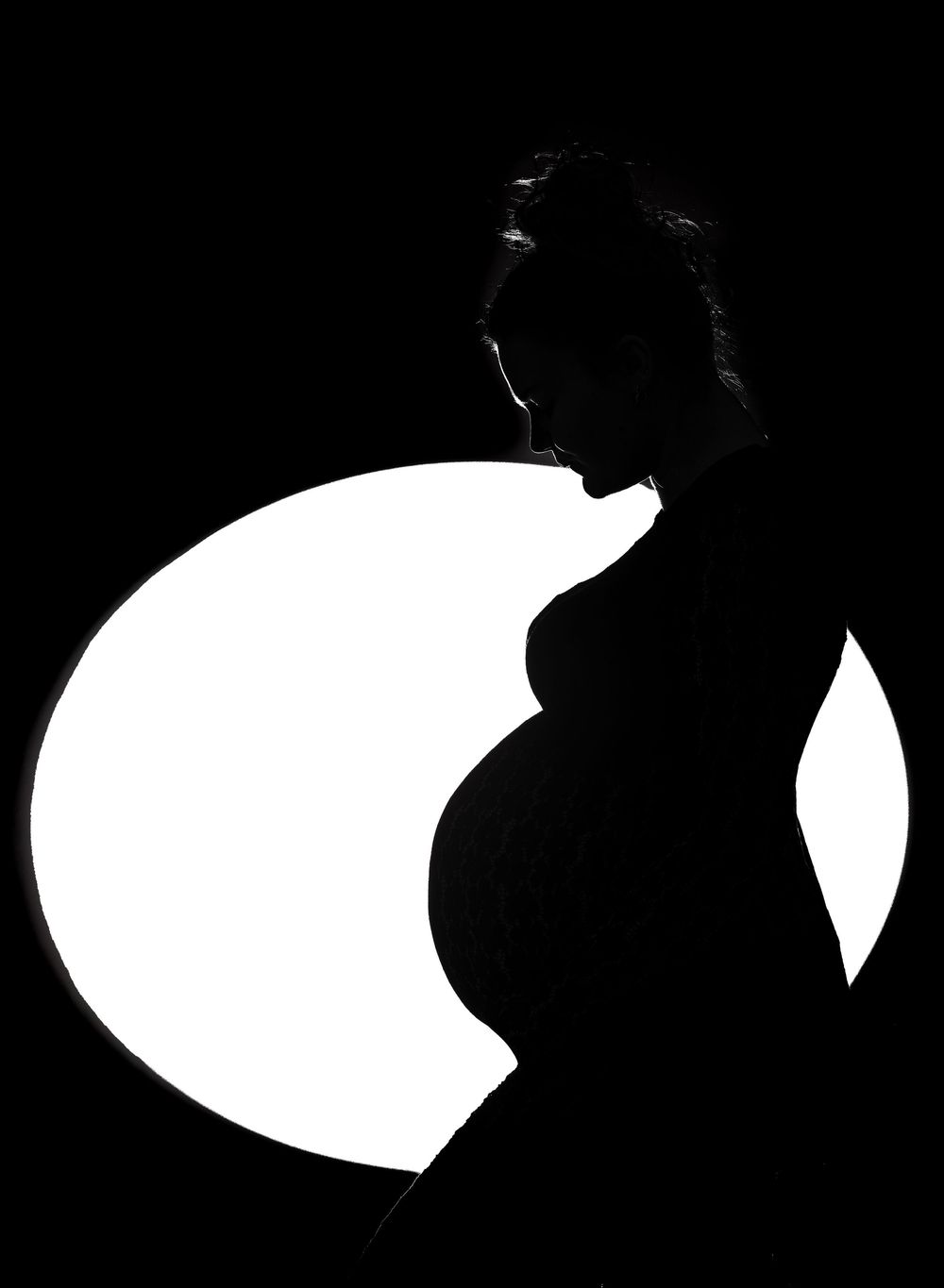 Silhouette of a pregnant woman in profile against a bright circular light on black background.