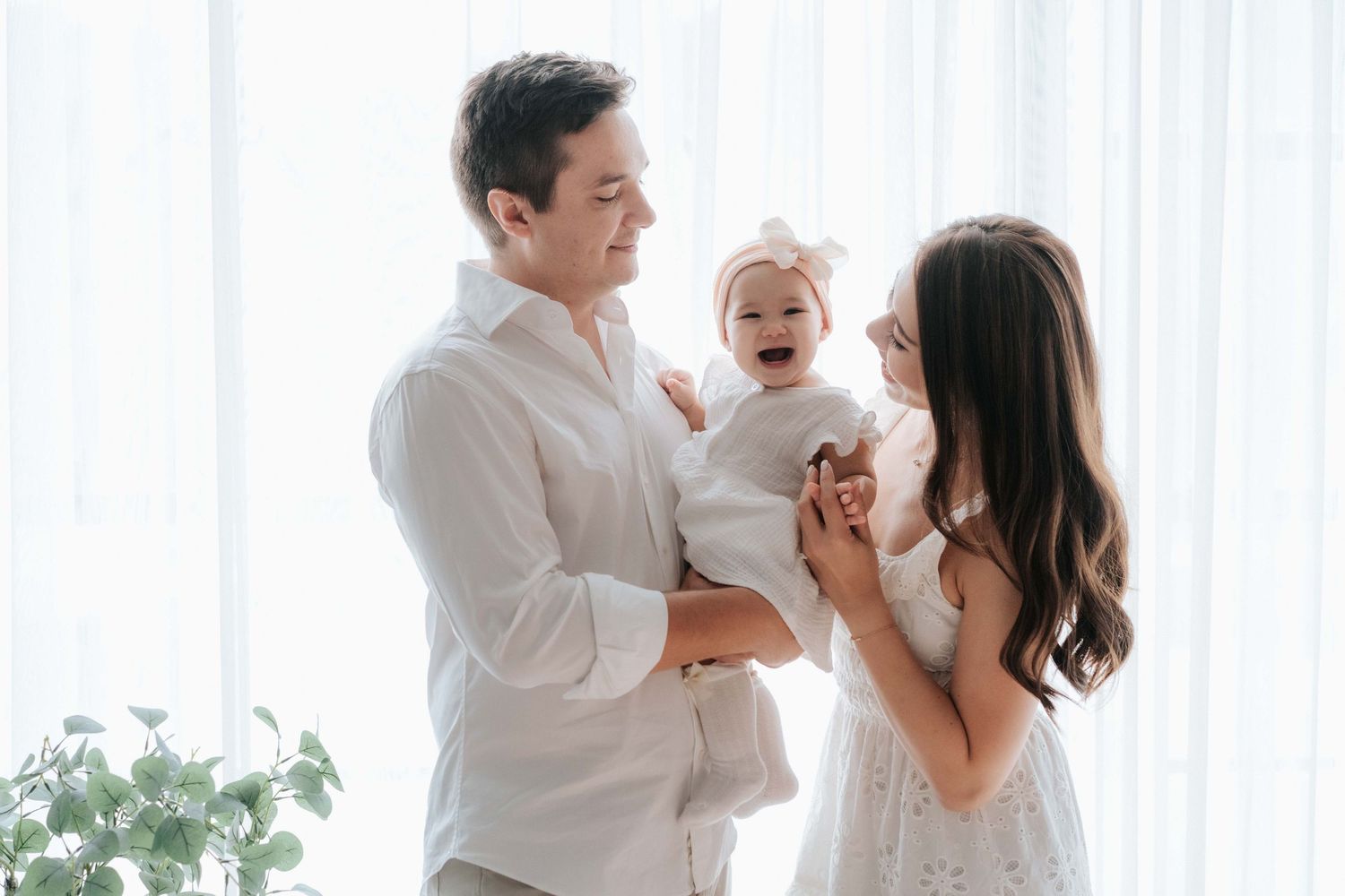 Happy family with mother and father in white outfits holding their laughing baby girl near bright curtained window.幸福的一家人，父母穿著白色服裝，抱著他們咯咯笑著的