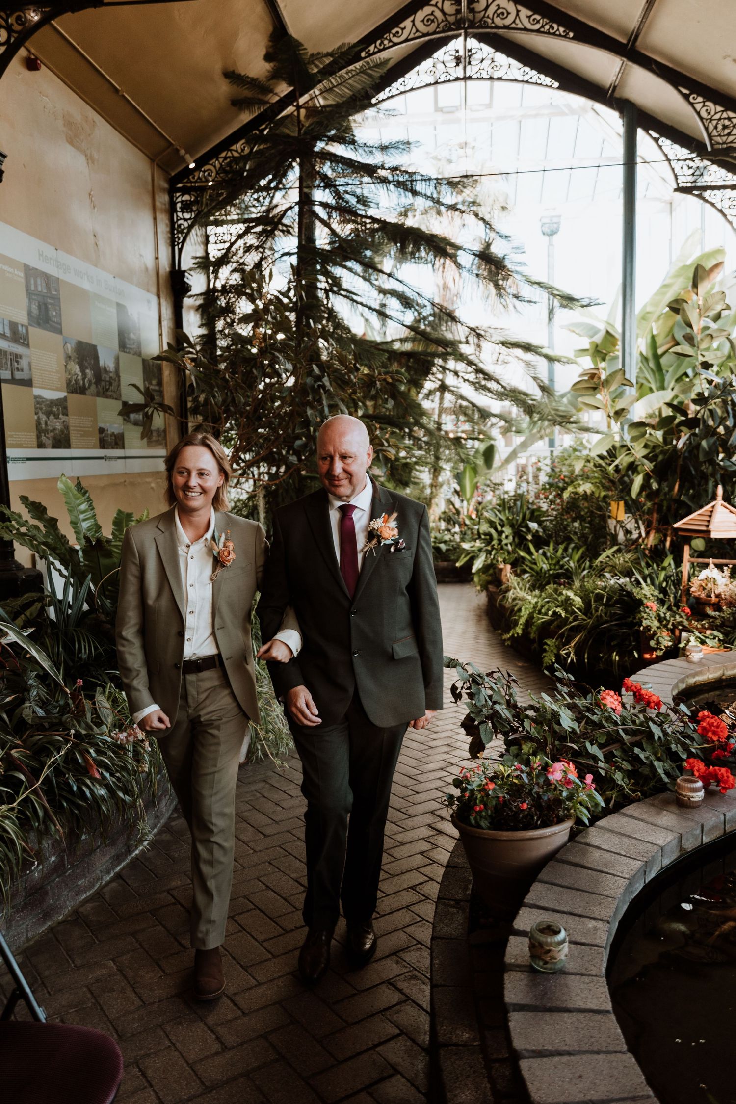 One half of the couple walking down the aisle with their dad and share a laugh while strolling through a greenhouse conservatory.