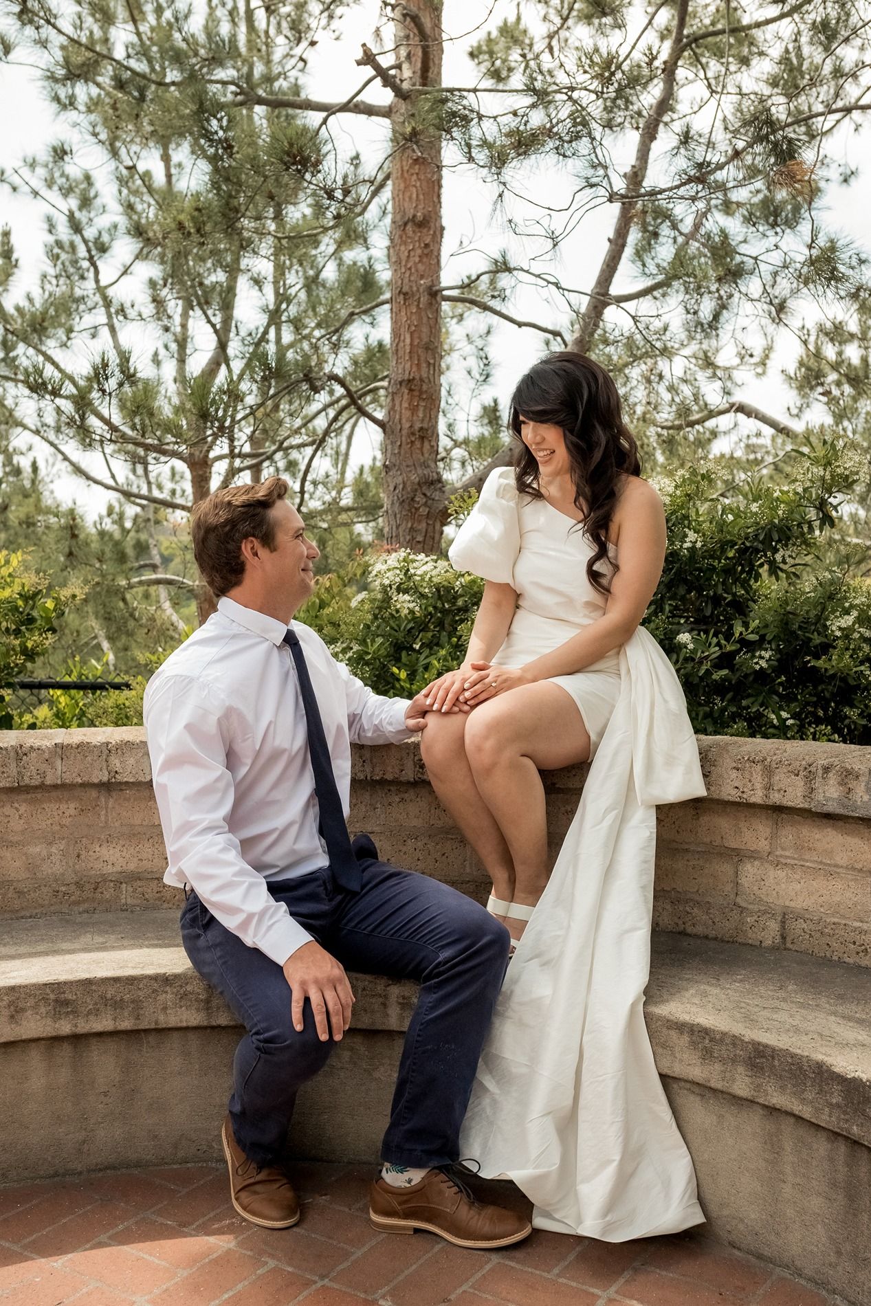 A couple in wedding attire shares a romantic moment on stone steps surrounded by pine trees in an outdoor setting at Balboa Park.
