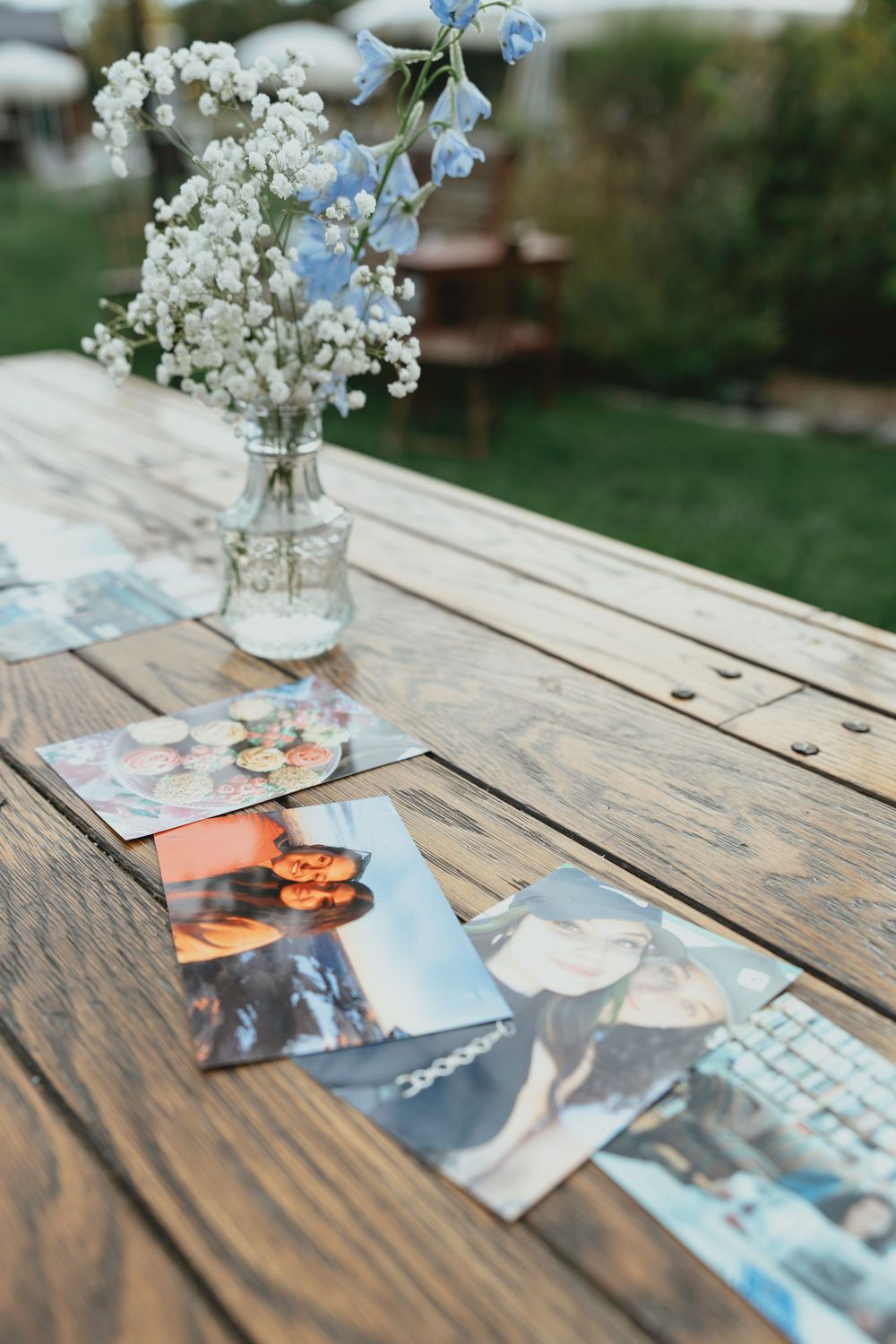 White flowers in a glass vase sit on a rustic wooden table with scattered photographs in an outdoor setting.