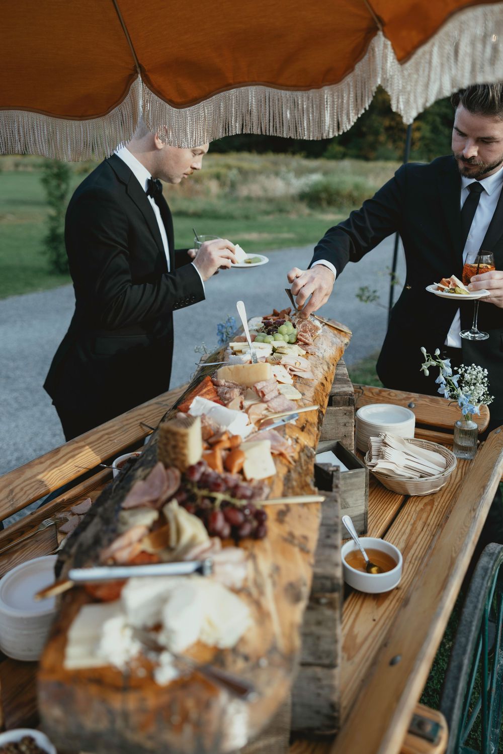 Guests serve themselves from an elegant charcuterie and appetizer spread on a rustic wooden table at an outdoor event.