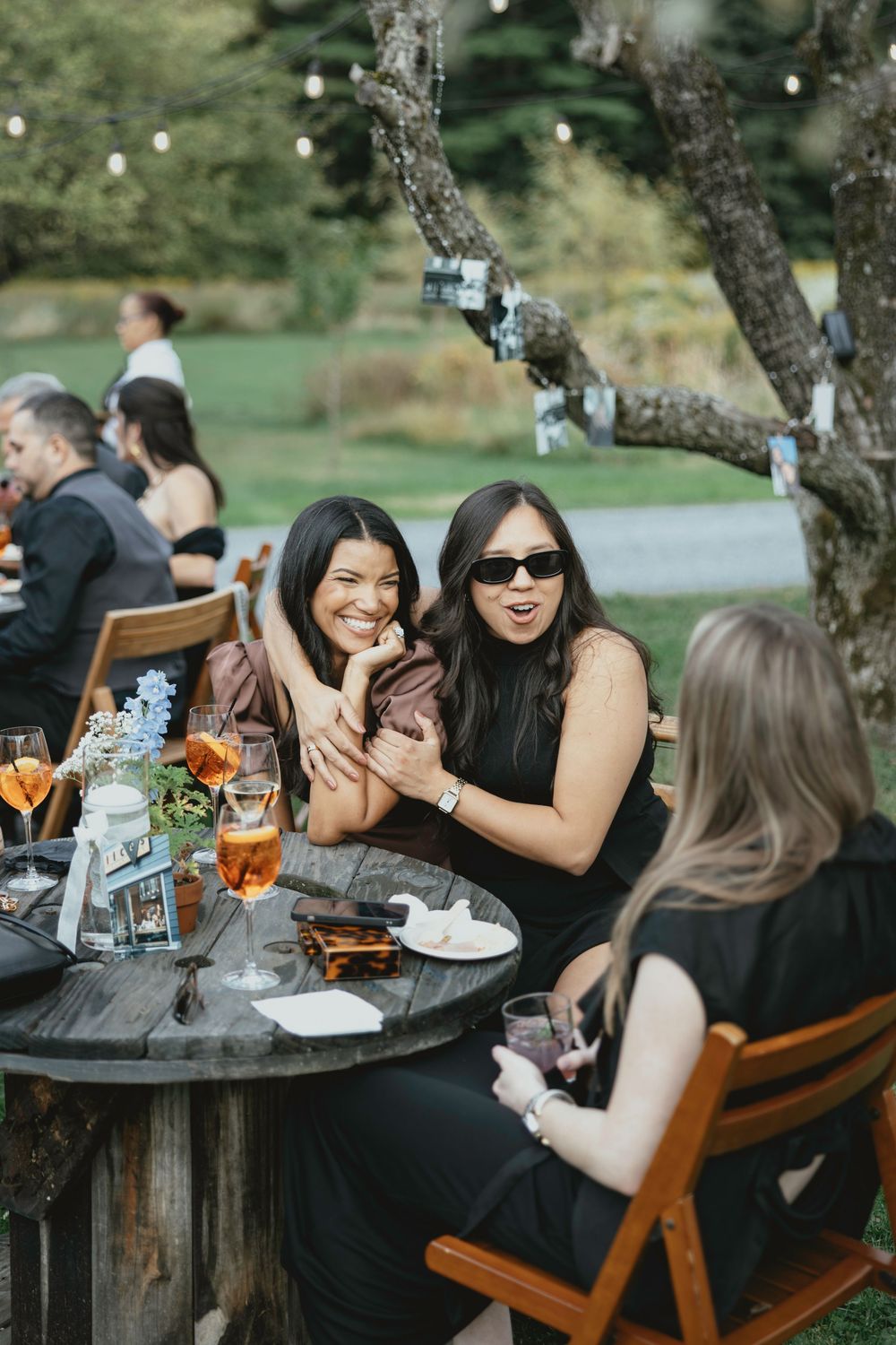 A group of friends sharing laughter and conversation while dining outdoors at a rustic wooden table.