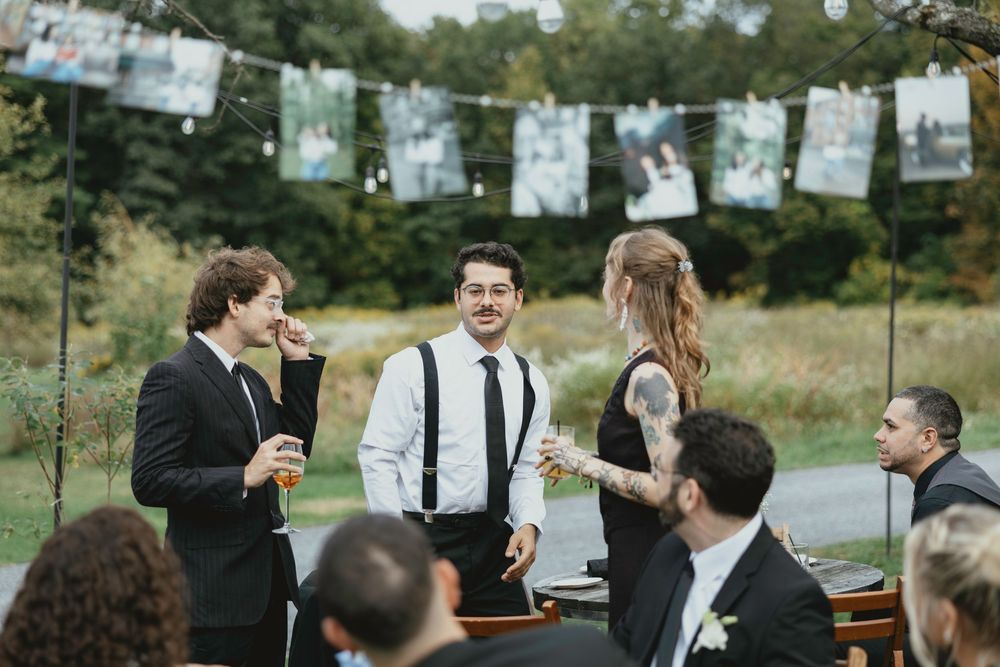 Wedding guests mingle and socialize during an outdoor reception with photos hanging on string lights in the background.
