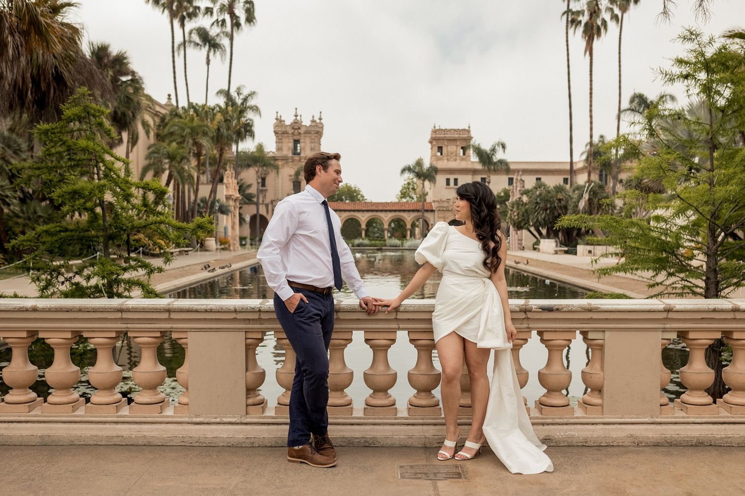 Couple in formal attire poses together on stone balcony with palm trees and historic architecture in background at Balboa Park in San Diego.