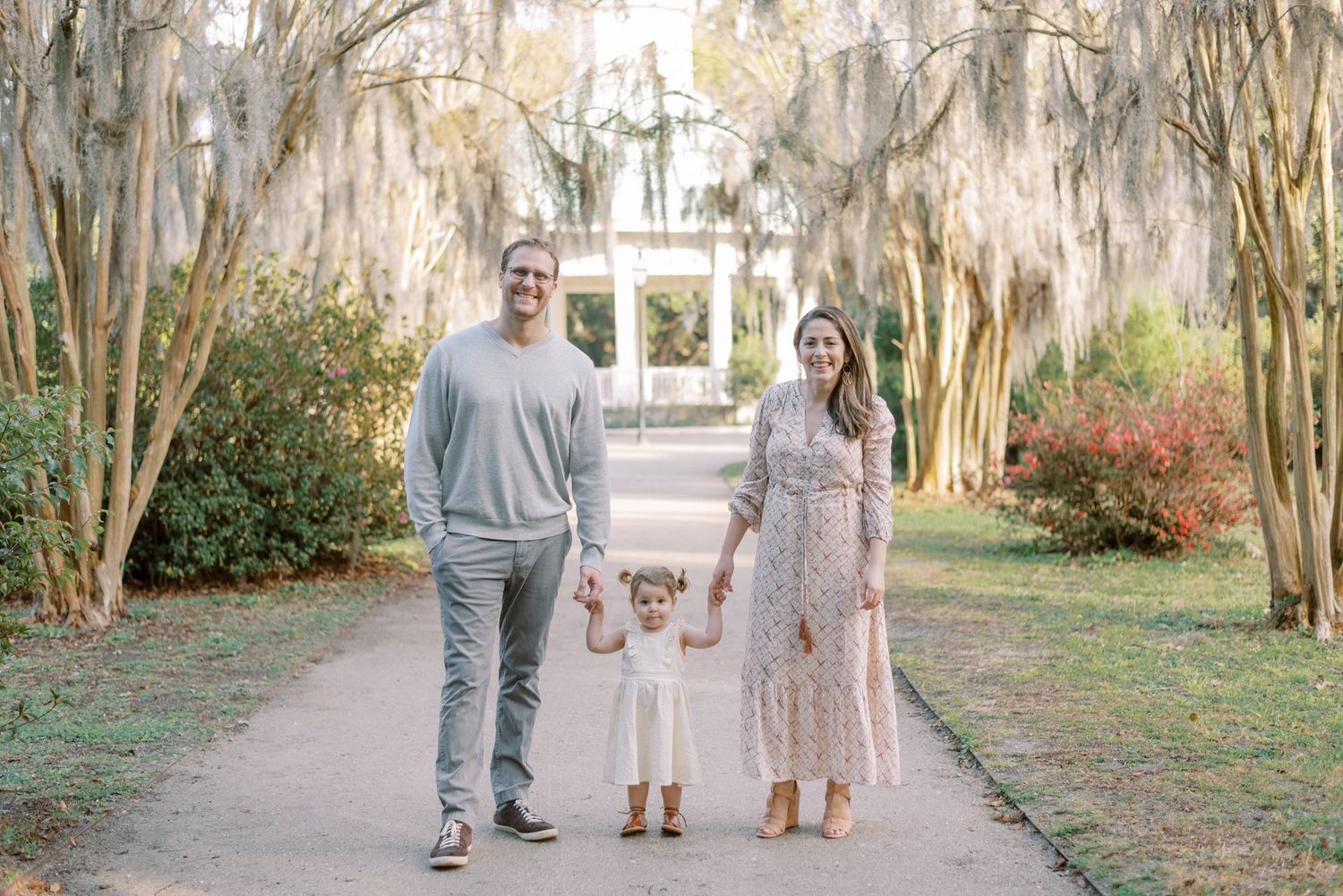 A family walks down a garden path lined with Spanish moss-draped trees and blooming azaleas in a Southern setting.