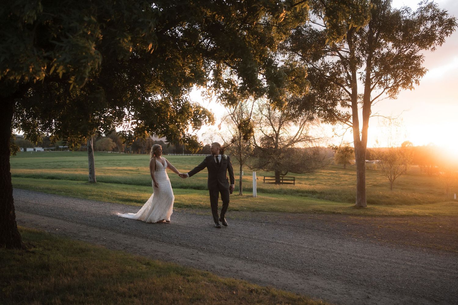 Two people in white and black outfits hold hands while walking through a sunlit forest path at dusk.