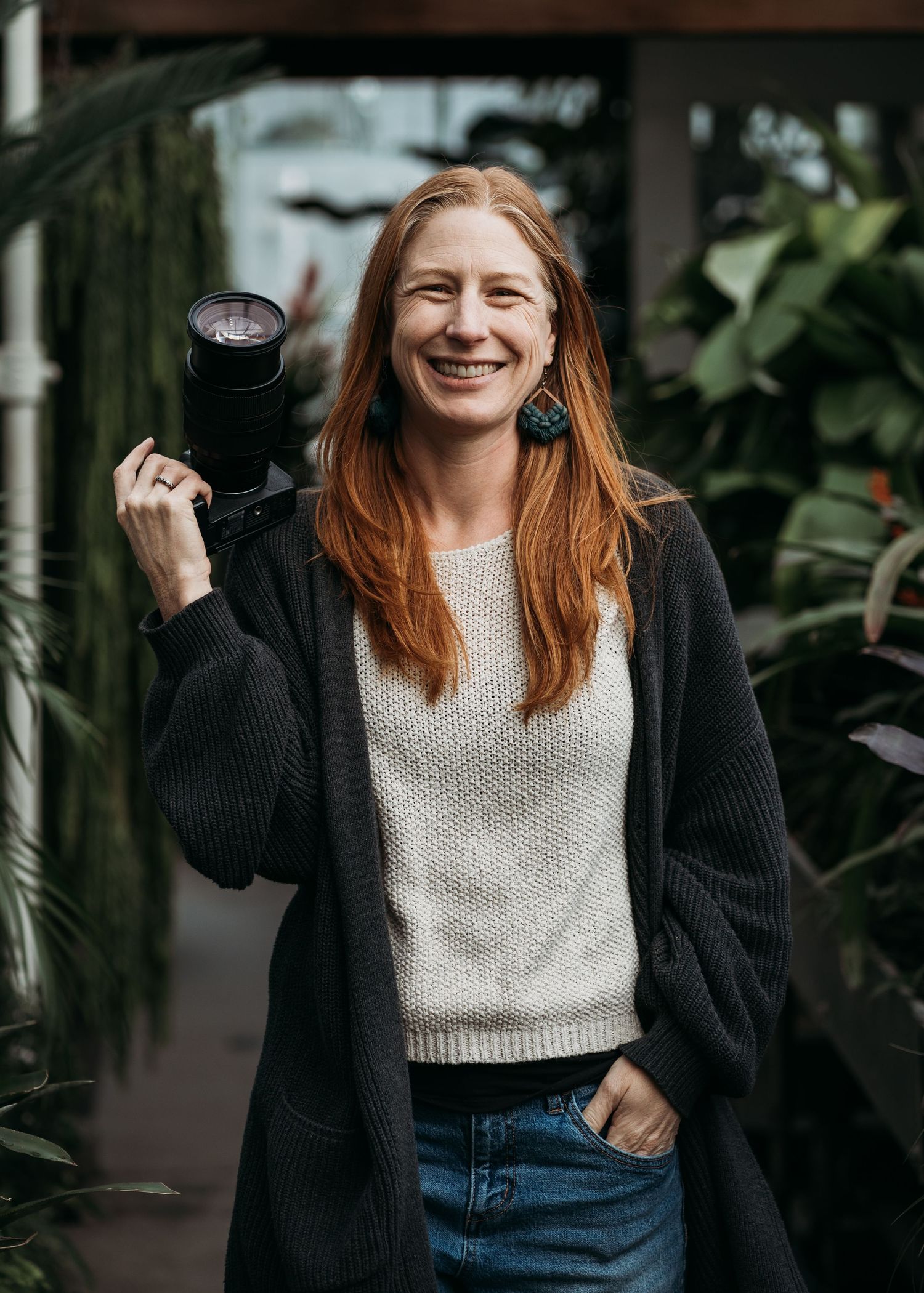 A person in a black cardigan and white sweater laughs joyfully while walking through an indoor space with plants.