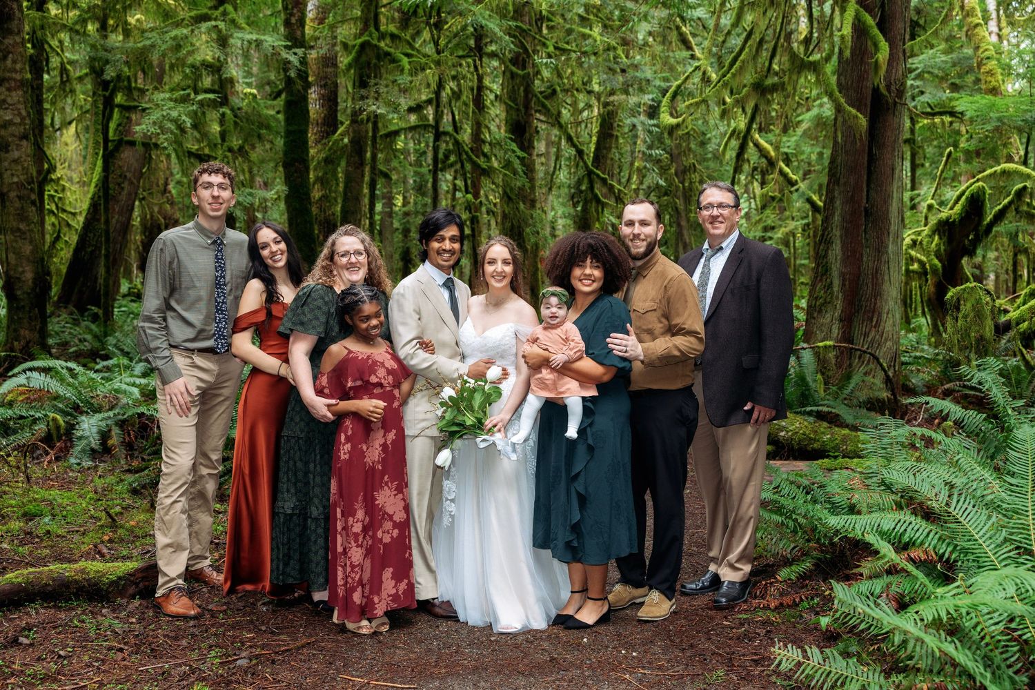 Wedding party stands together in lush green forest setting surrounded by tall trees and ferns.