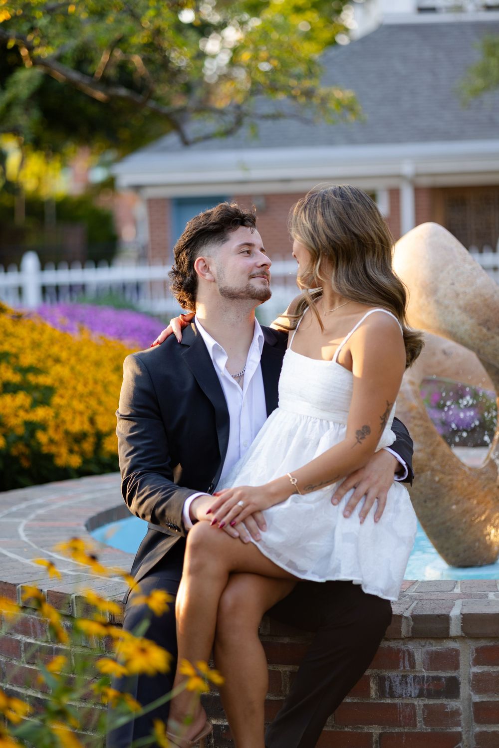 Couple sitting together on a fountain ledge surrounded by colorful flowers, gazing at each other lovingly.
