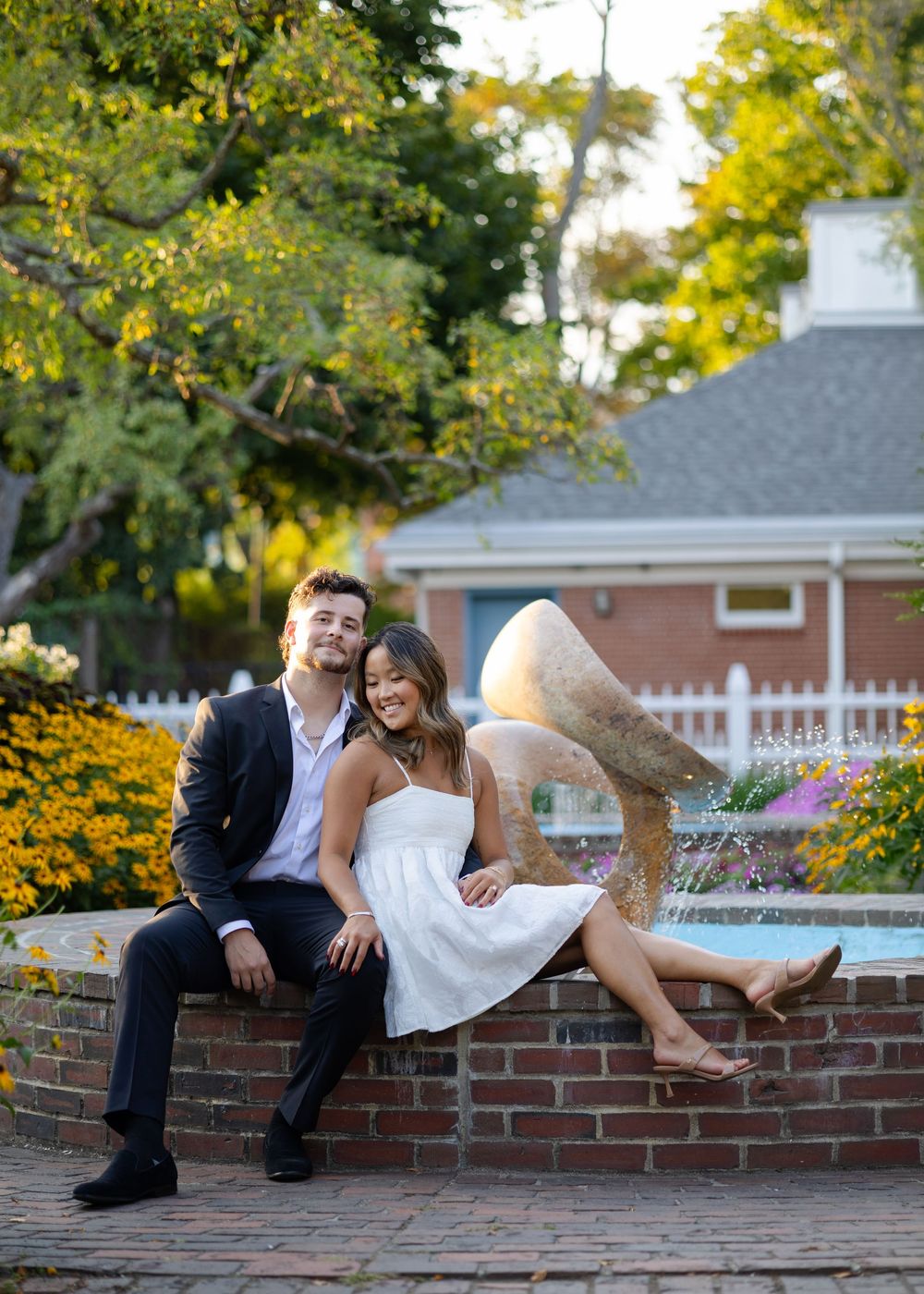 Smiling engaged couple sitting by a garden fountain with yellow flowers and lush trees in background.