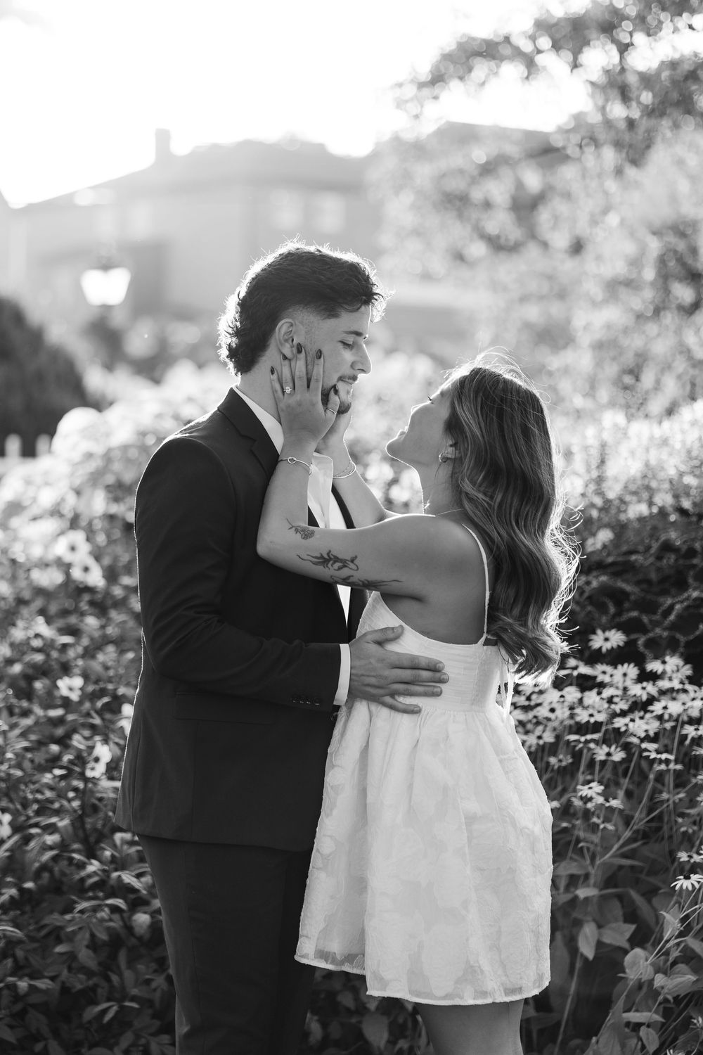 Black and white photo of a couple embracing in a sunlit garden with flowers.