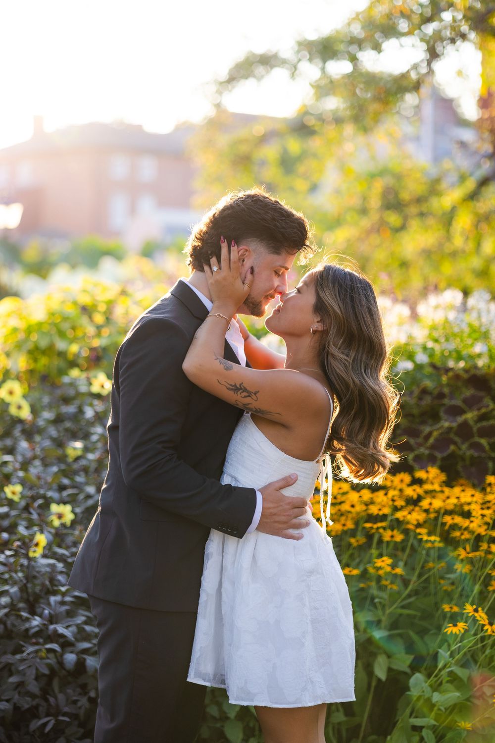 Couple sharing romantic kiss in golden sunset light surrounded by vibrant yellow garden flowers.