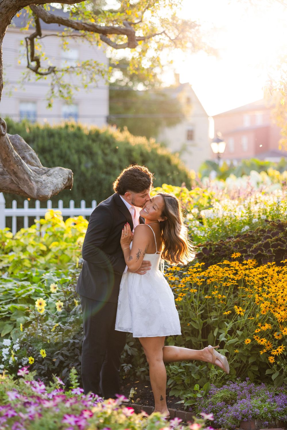 Couple sharing a romantic kiss in a sunny garden with colorful flowers, woman in white dress lifting her foot.
