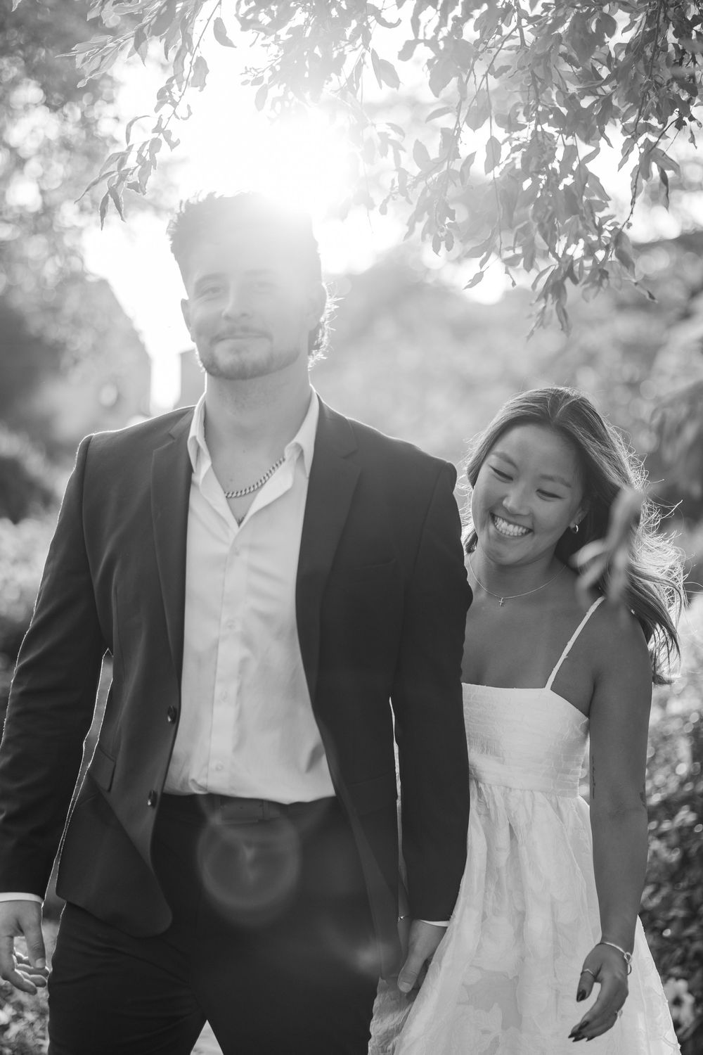 Couple posing outdoors in black and white, woman in white dress smiling, man in dark blazer.