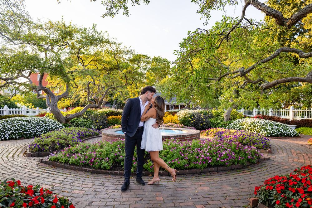 Couple sharing a tender kiss surrounded by colorful flower gardens and lush green trees in a scenic park setting.