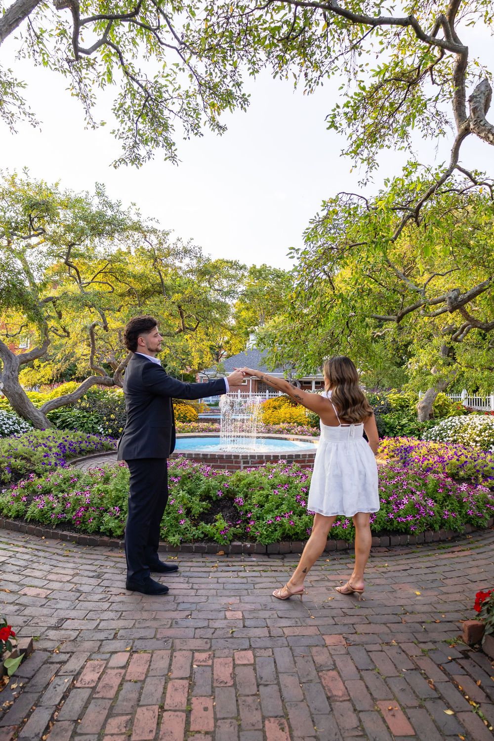 Couple dancing together in a lush garden with a fountain and colorful flowers in the background.