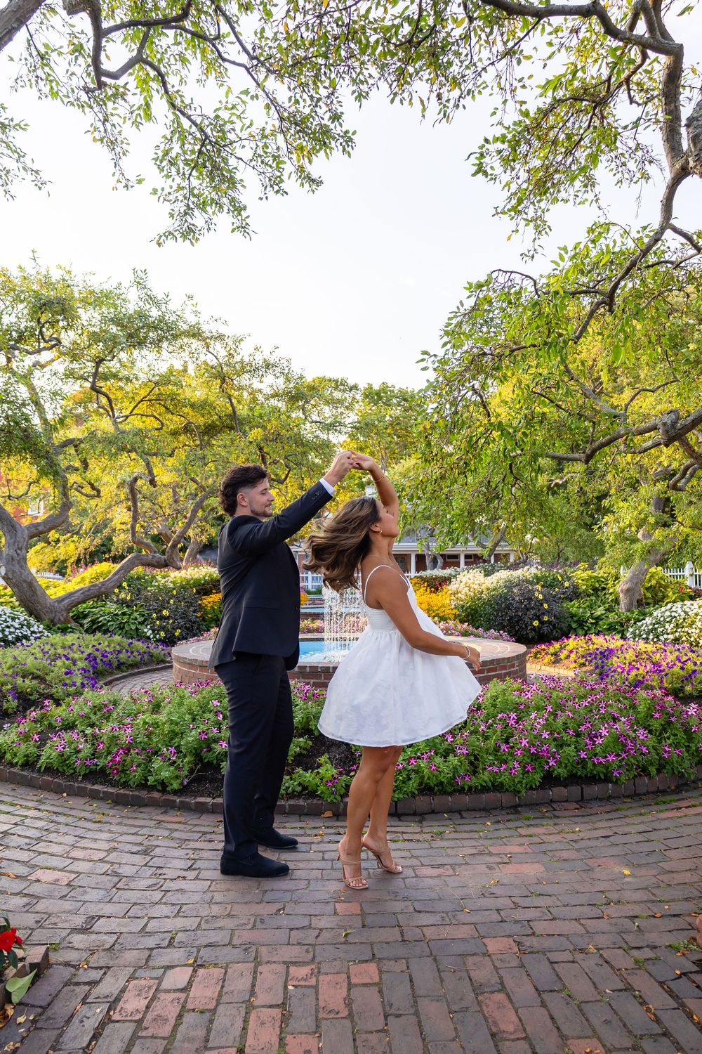 Couple dancing and twirling in white dress on brick path in colorful formal garden setting.