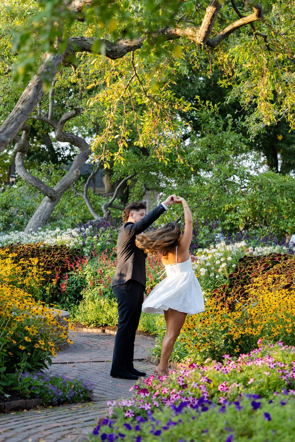 A couple dancing gracefully on a garden path surrounded by colorful blooming flowers and lush green trees.