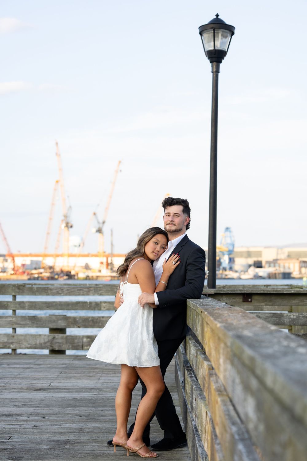 Couple in formal wear embracing on a waterfront pier, woman in white dress, man in black suit, with harbor cranes in background.