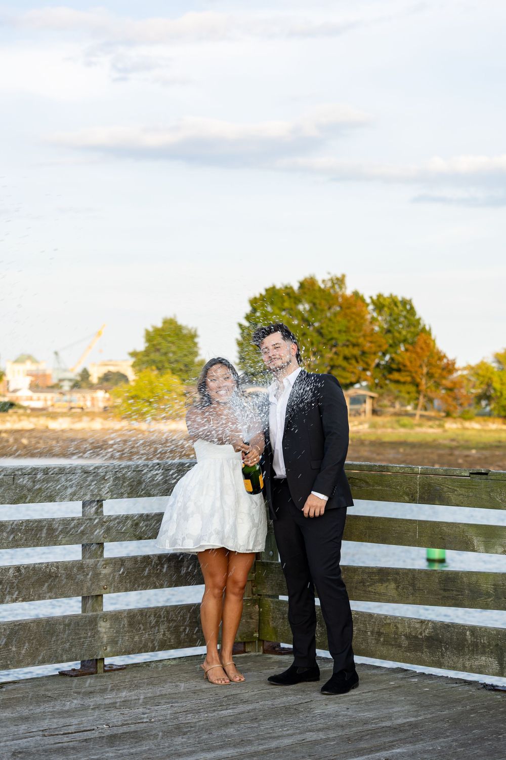 Newlyweds celebrate on a waterfront pier by popping champagne, bride in white dress and groom in black suit.
